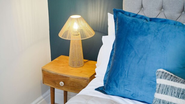 Ein Bett mit weißer Decke und blauem Kopfkissen, ein Nachttisch aus Holz mit einer Lampe und eine Wand mit dunkelblauer Wandverkleidung.