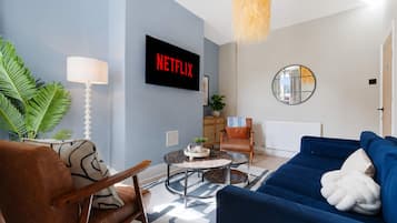 Un salón con un sofá azul, un sillón de cuero marrón, una mesa de centro, un televisor mostrando 'NETFLIX', un espejo redondo y una planta.