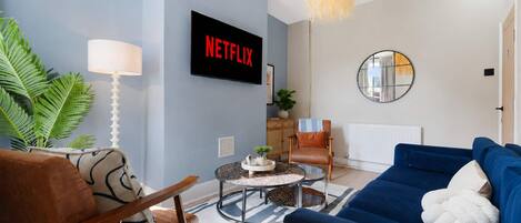 Een woonkamer met een blauwe bank, een bruine leren fauteuil, een salontafel, een televisie waarop 'NETFLIX' staat, een ronde spiegel en een plant.
