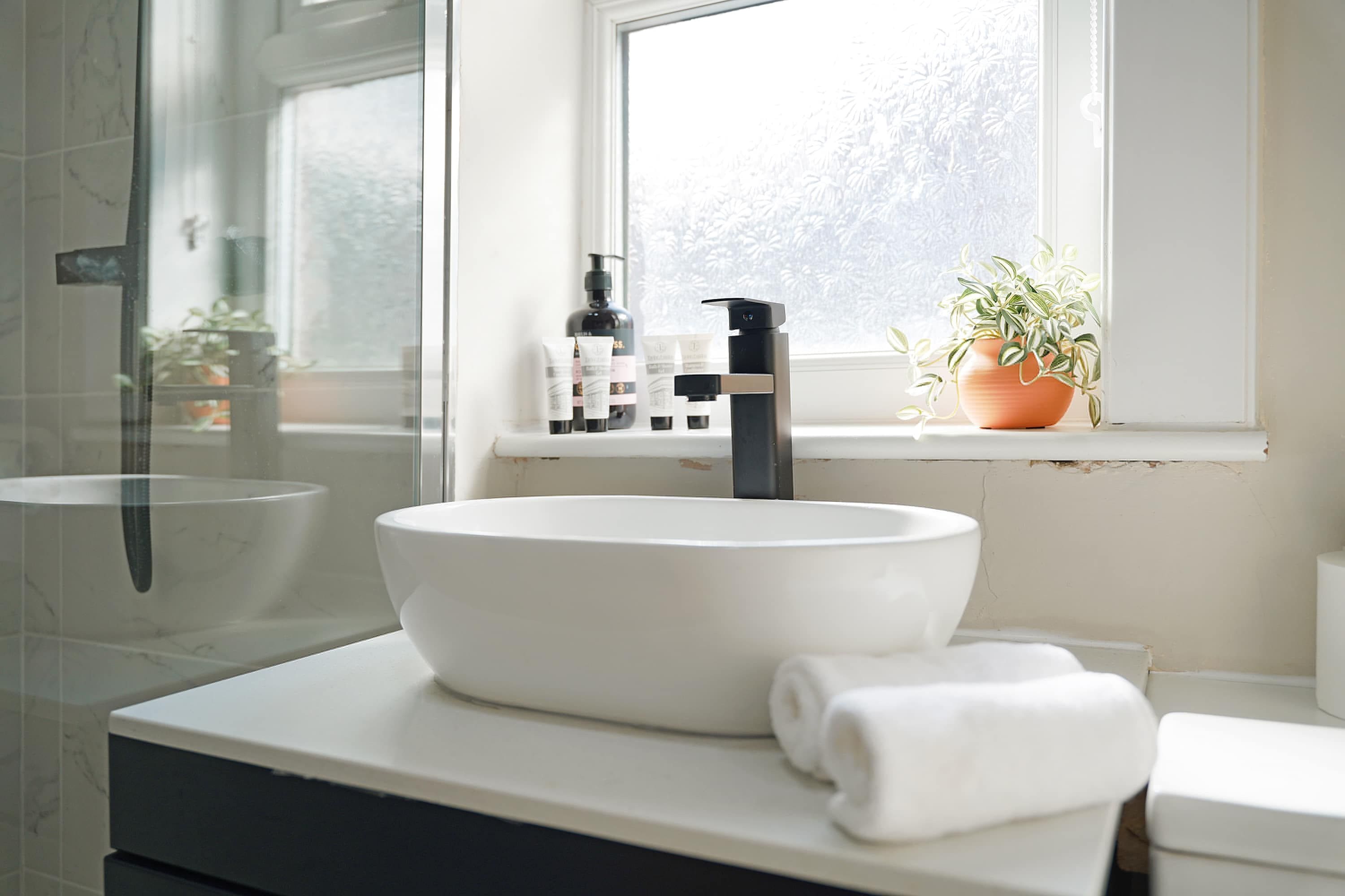 Ein Badezimmer mit einem weißen Waschbecken, einem schwarzen Wasserhahn und einem Fenster mit Blick auf Bäume.
