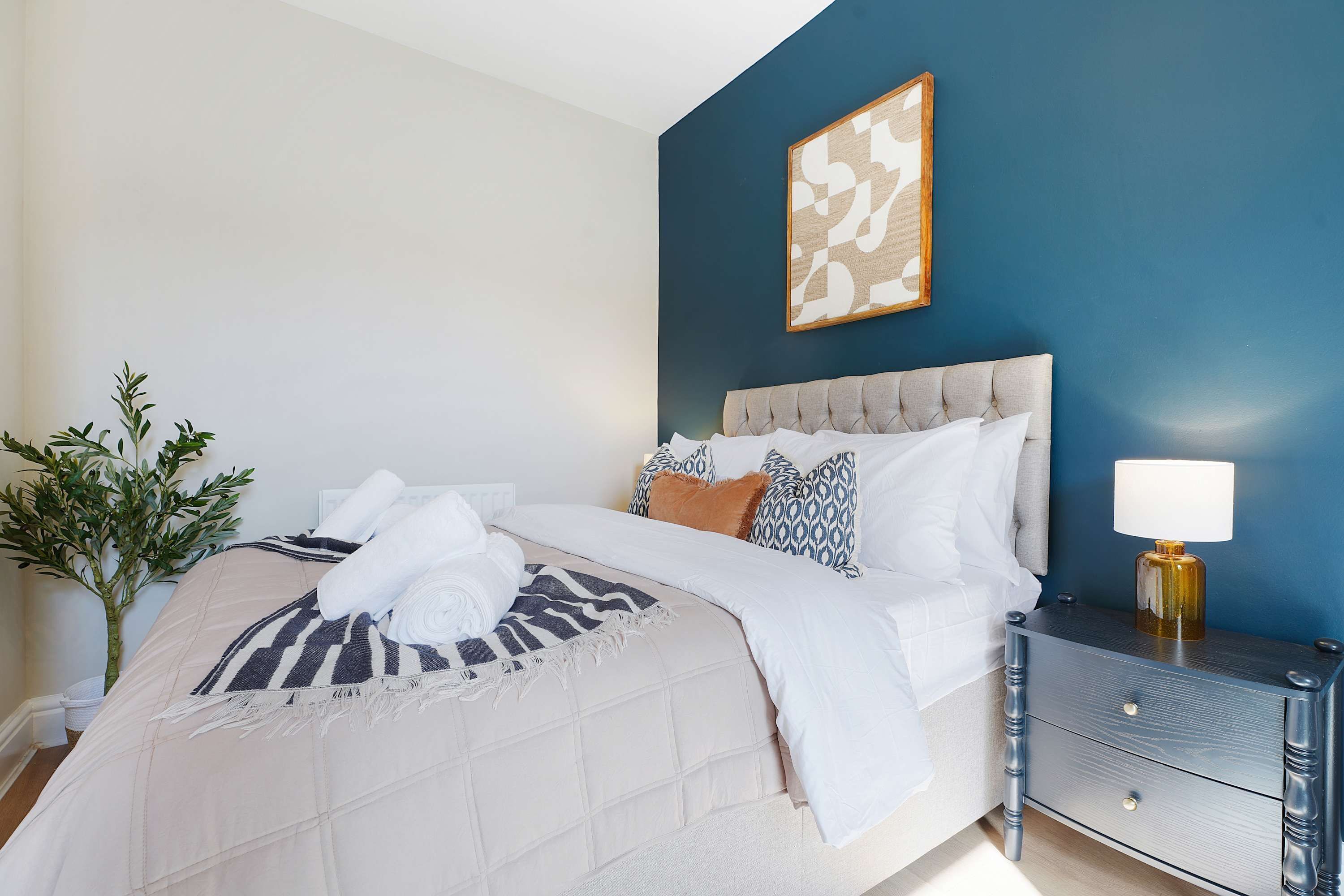 A neatly made bed with a striped blanket and rolled towels, a bedside table with a lamp, and a wall-mounted abstract art piece.