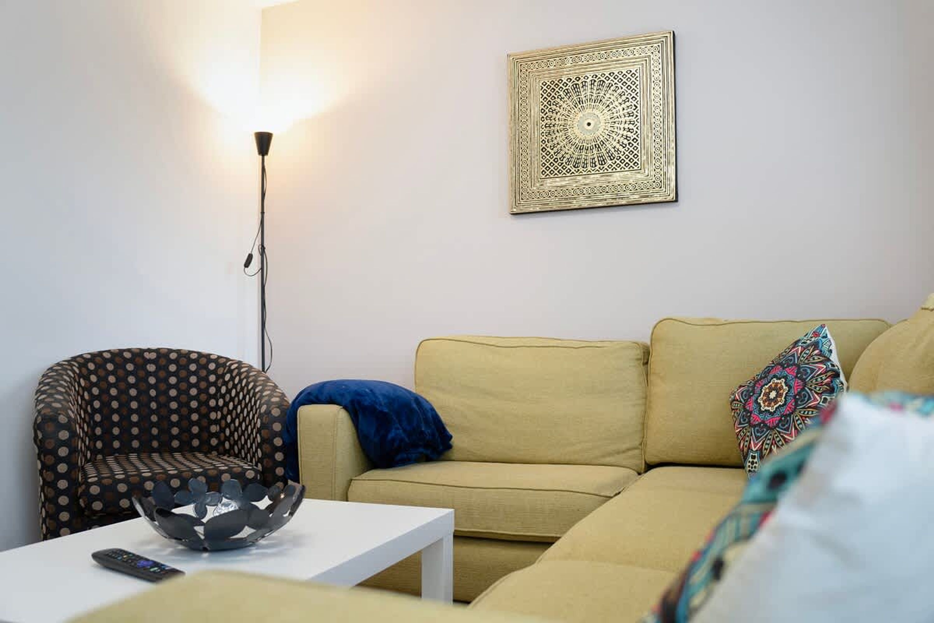 En stue med sofa, lænestol med mønster, hvidt sofabord, gulvlampe og en dekorativ vægdekoration.