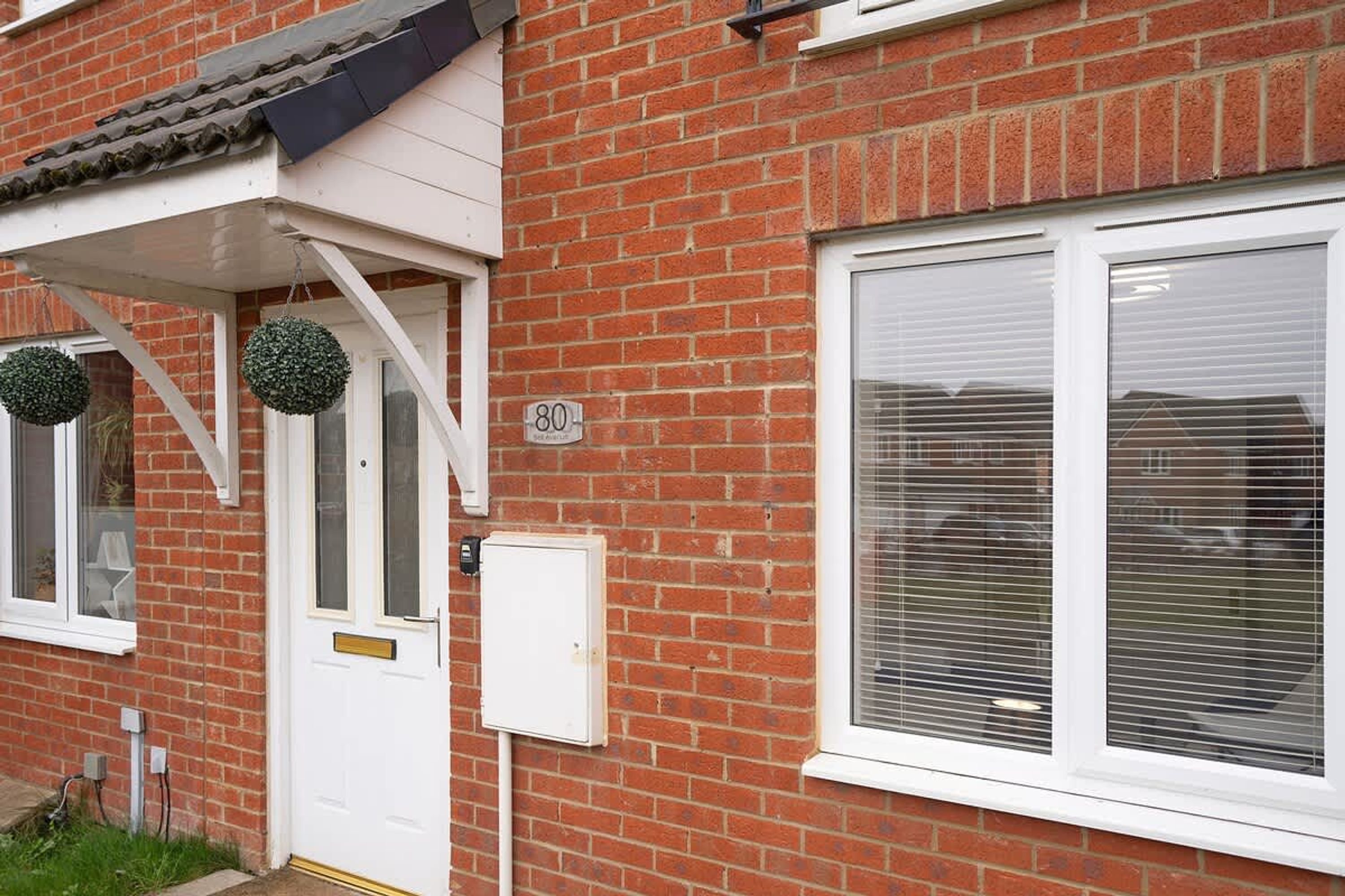 Une maison en briques, d’habitation, avec une porte blanche, une plaque portant le numéro « 80 » et une fenêtre avec des stores.