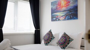 A hotel room with a bed, two decorative pillows, a painting on the wall, and a window with curtains. - Spacious Retreat for 10 Perfect for Families (Durham)
