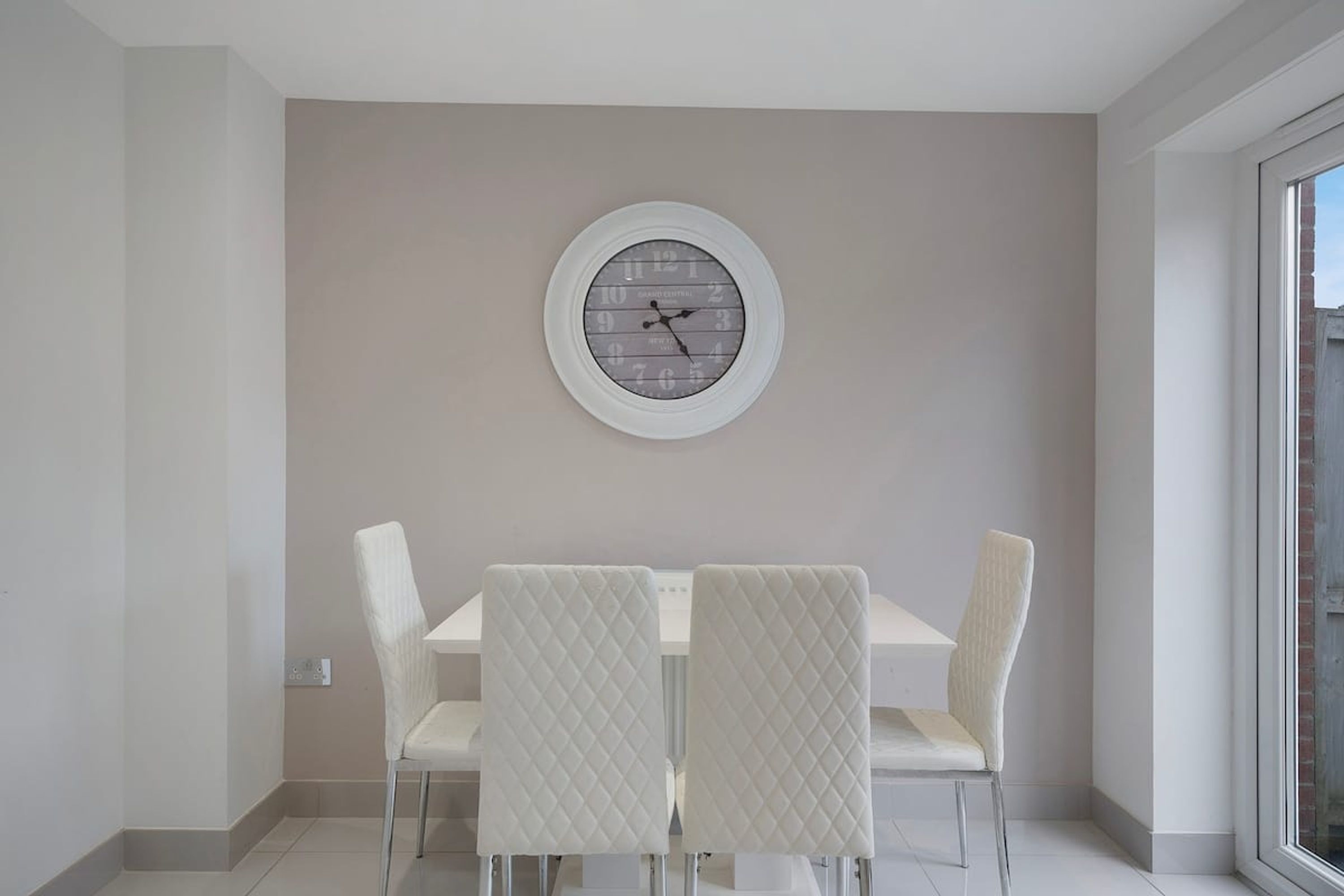 Un comedor con un reloj de pared redondo, sillas blancas y un ventanal grande.