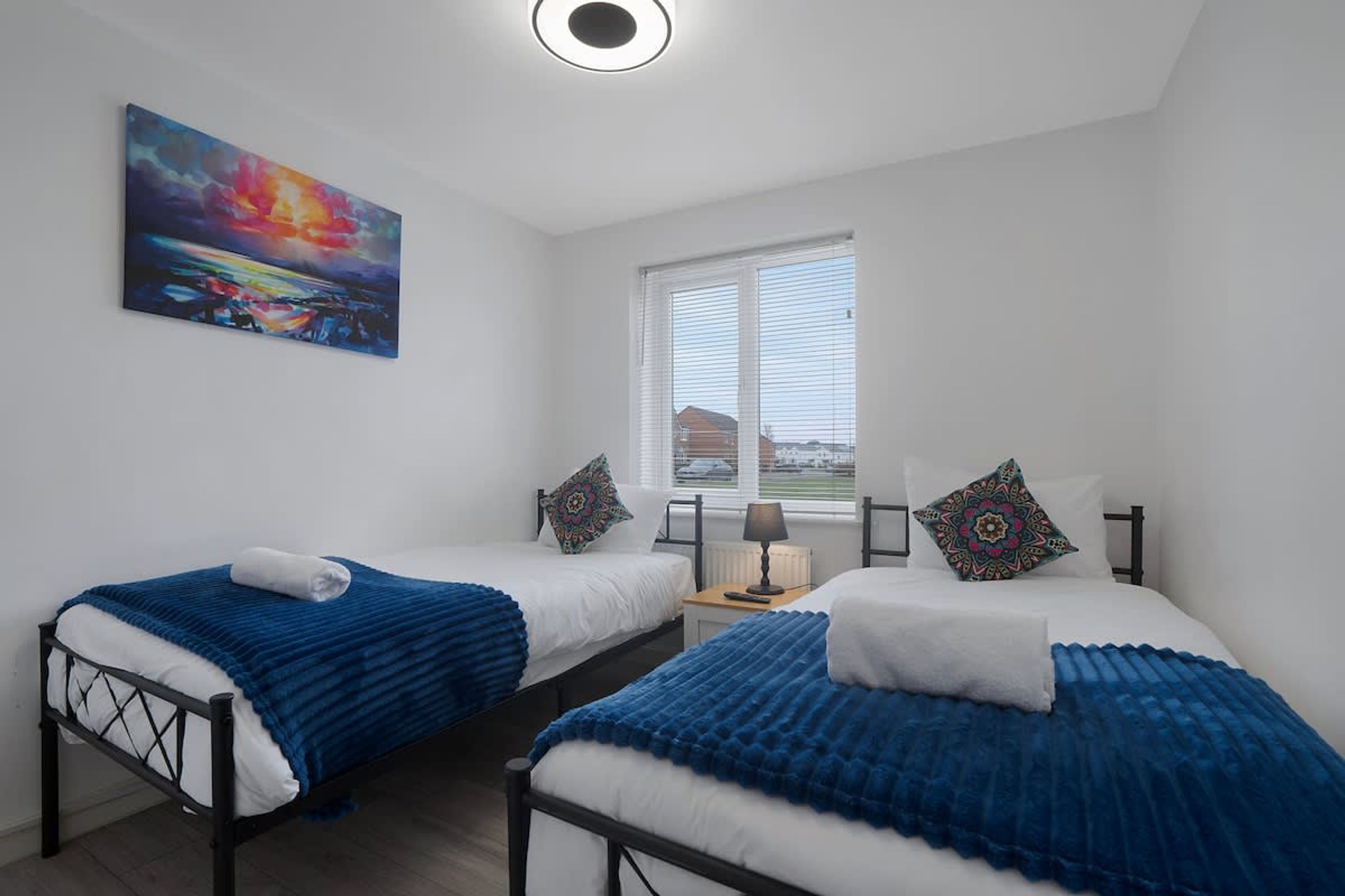 A room with two beds, a painting on the wall, and a window with blinds.