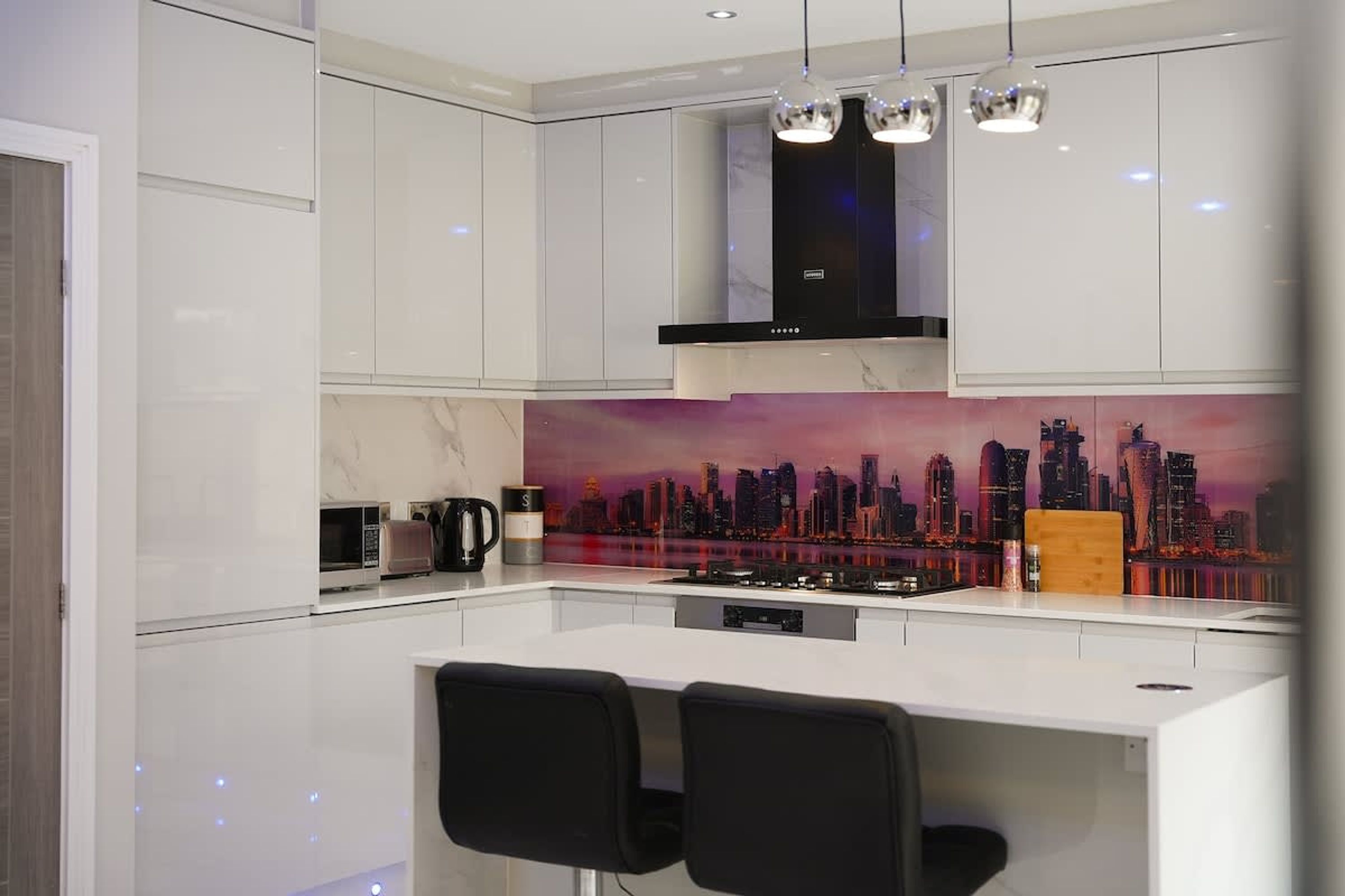 Une cuisine moderne avec des meubles blancs, une hotte noire et une fresque représentant un paysage urbain sur le mur derrière l’évier.