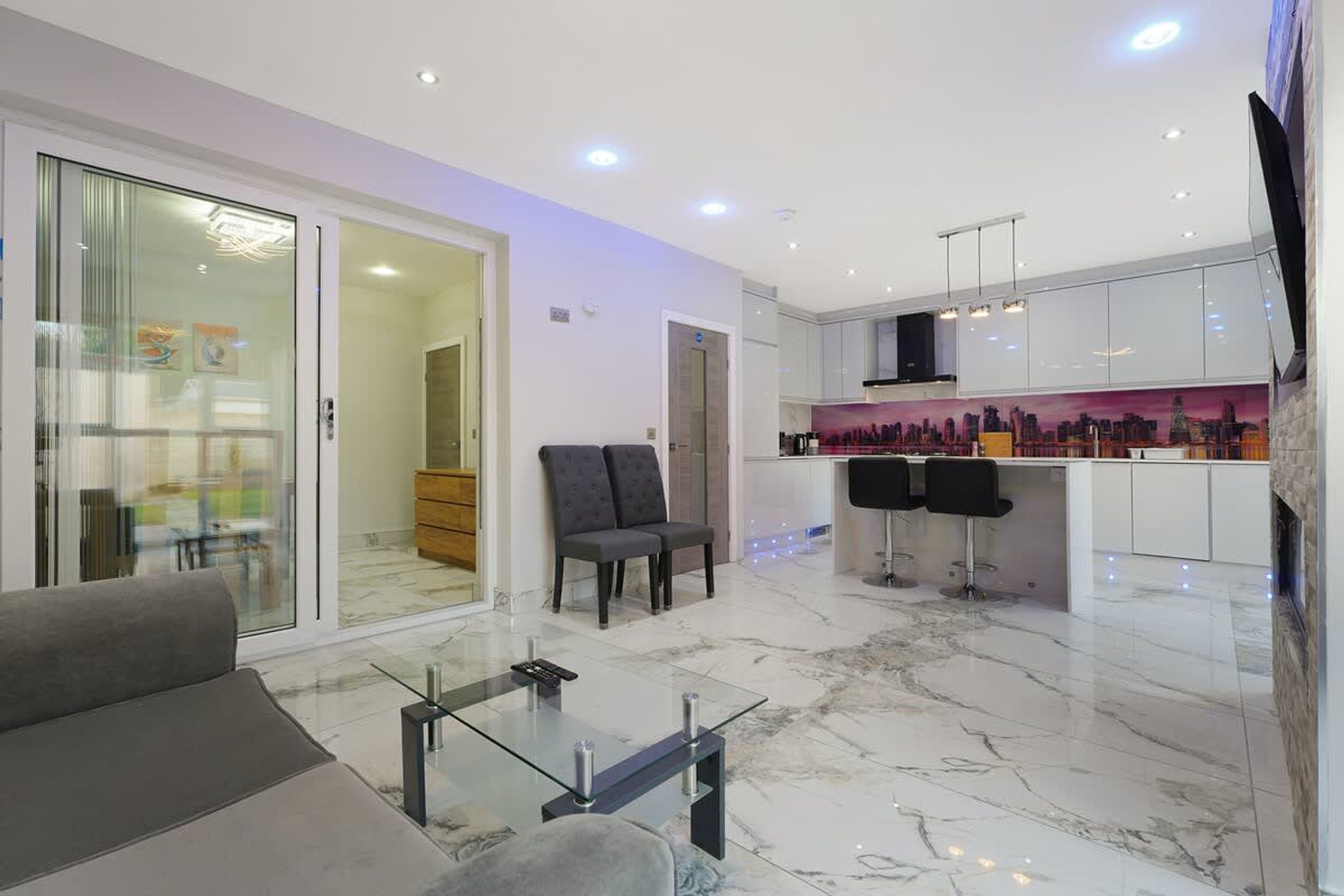 A modern living room with a glass coffee table, a sofa, and a kitchen area with a bar and seating.