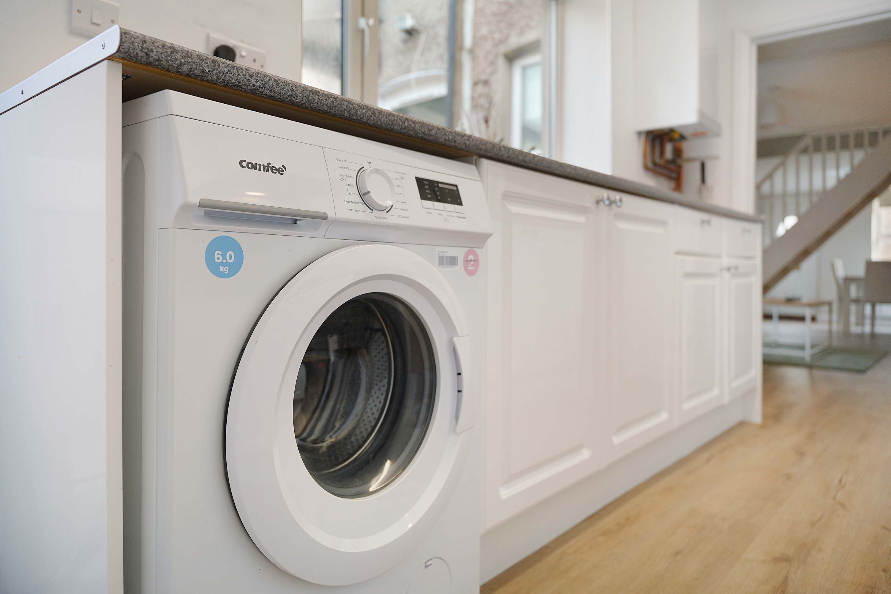 A front-loading Comfee washing machine with a 6.0 kg capacity, located in a modern kitchen with wooden flooring and white cabinetry.