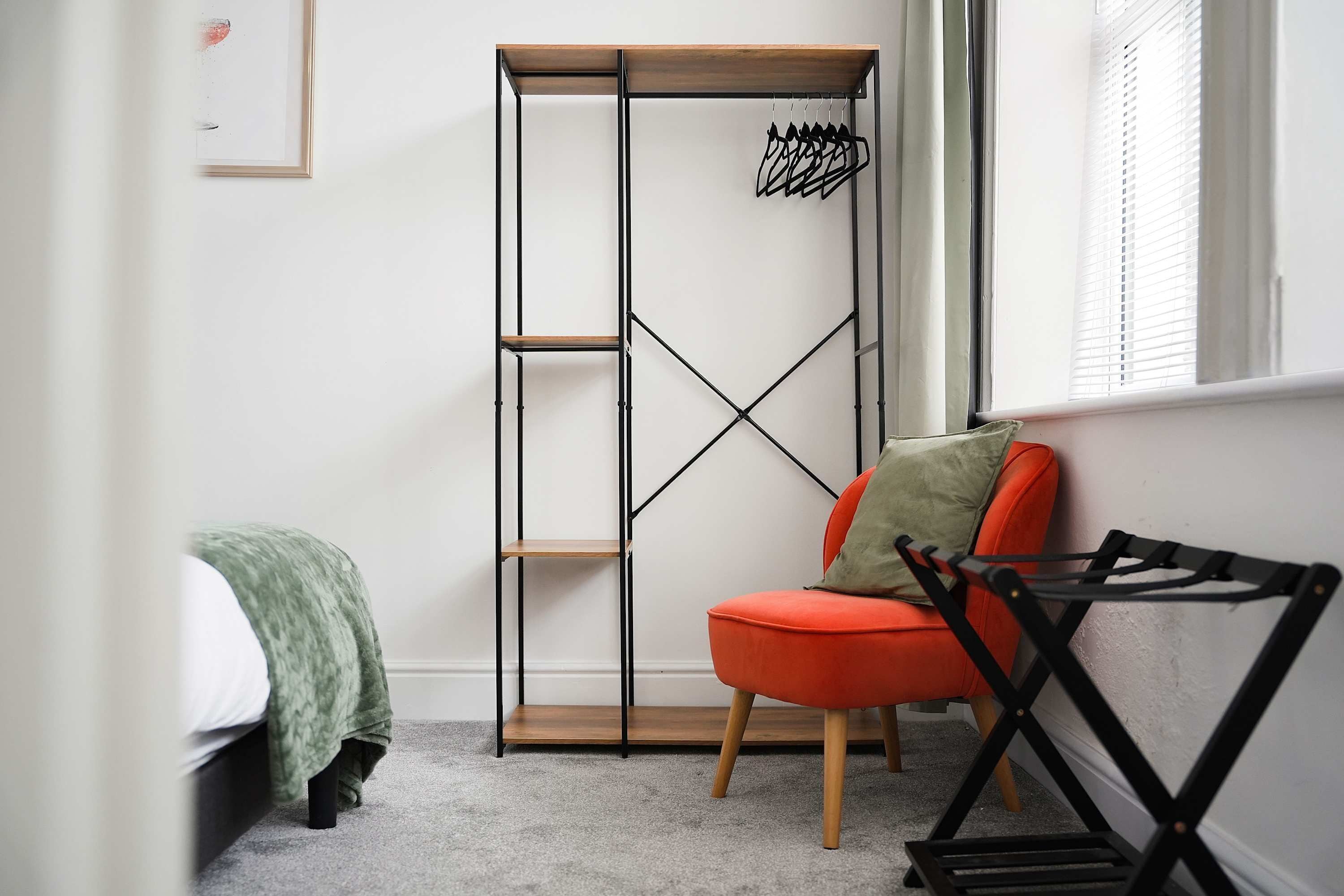 A room with a bed, a green blanket, a red chair, a wooden wardrobe, and a black hanger rack.