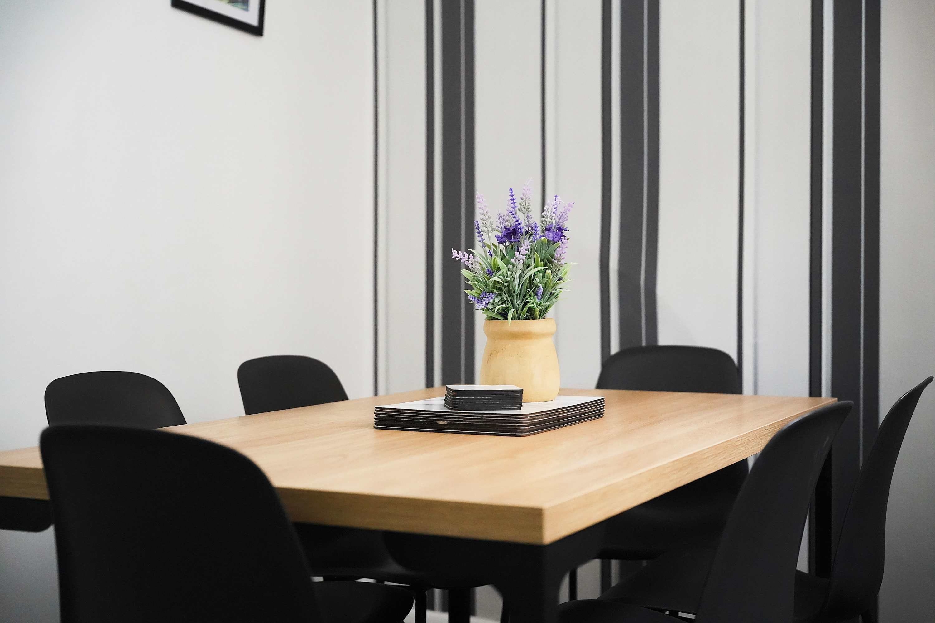 Een vergaderruimte met een houten tafel, zwarte stoelen en een potplant met paarse bloemen.