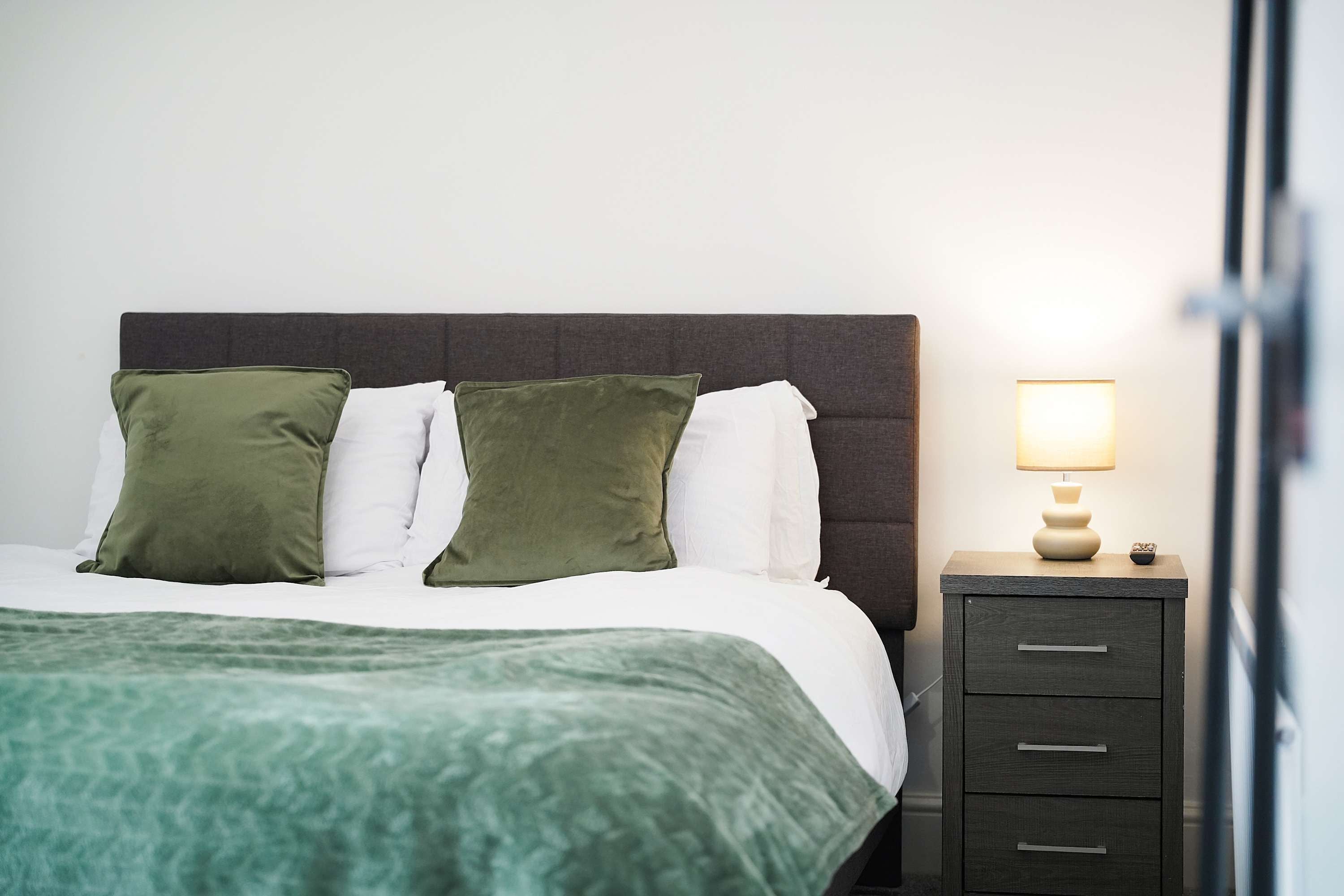 Ein ordentlich bezogenes Bett mit grüner Decke, einer Nachttischlampe und einem dunklen Nachttisch mit Schubladen.