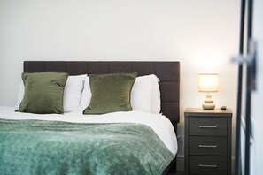 A neatly made bed with a green blanket, a bedside lamp, and a dark-colored nightstand with drawers.