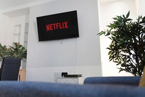A living room with a mounted television displaying 'NETFLIX' in red letters, a gaming console, and a potted plant. - 4BR on Durham Road Perfect For Large Groups (Stanley)
