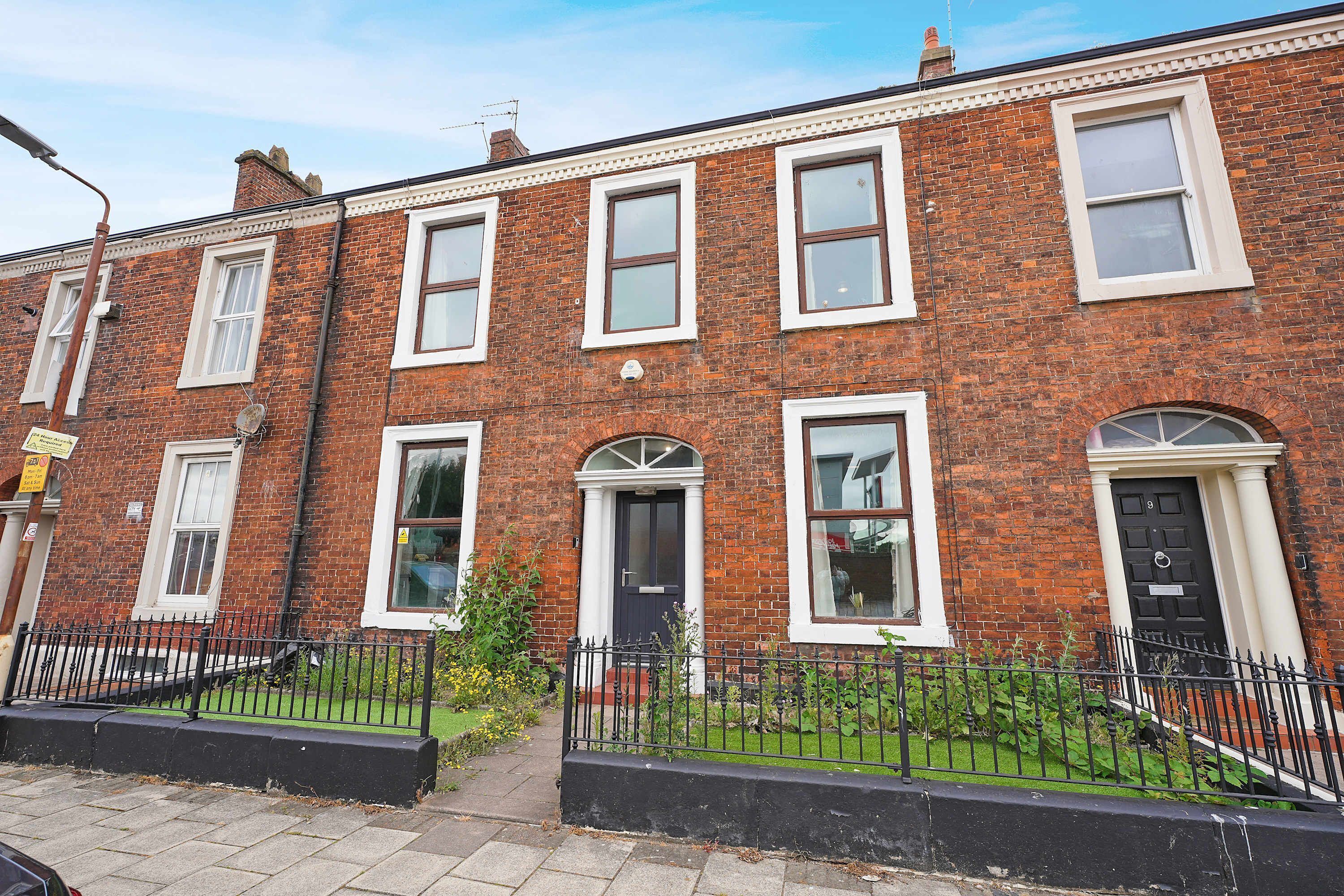 A two-story brick terraced house with white-framed windows, a black front door, and a black metal fence.