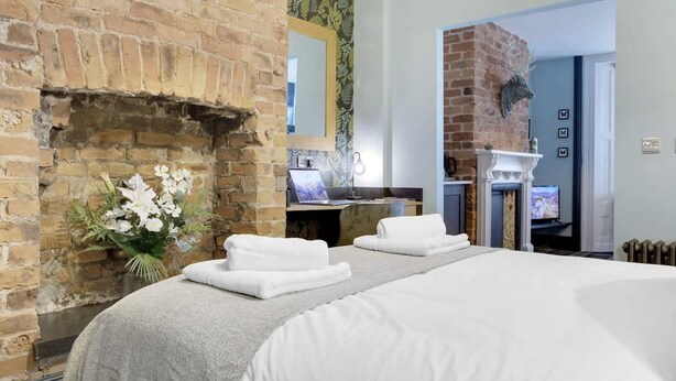 A hotel room with a brick fireplace, a bed with white linens, a small table with a laptop, and a television. - Stylish 1BR Home Near Station by Stay With Us (Hartlepool)