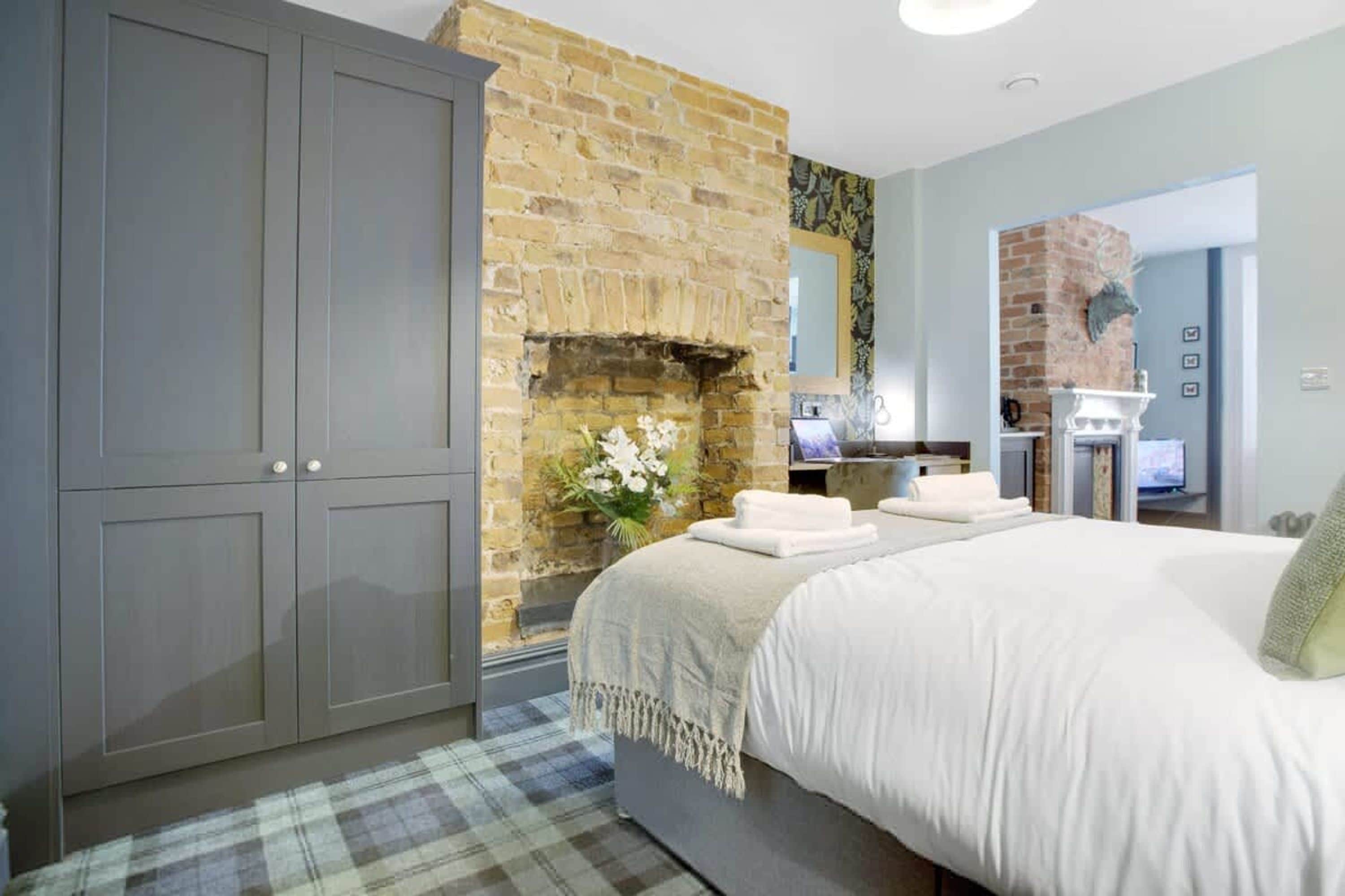 A bedroom with a large bed, grey wardrobe, brick wall, and a fireplace.