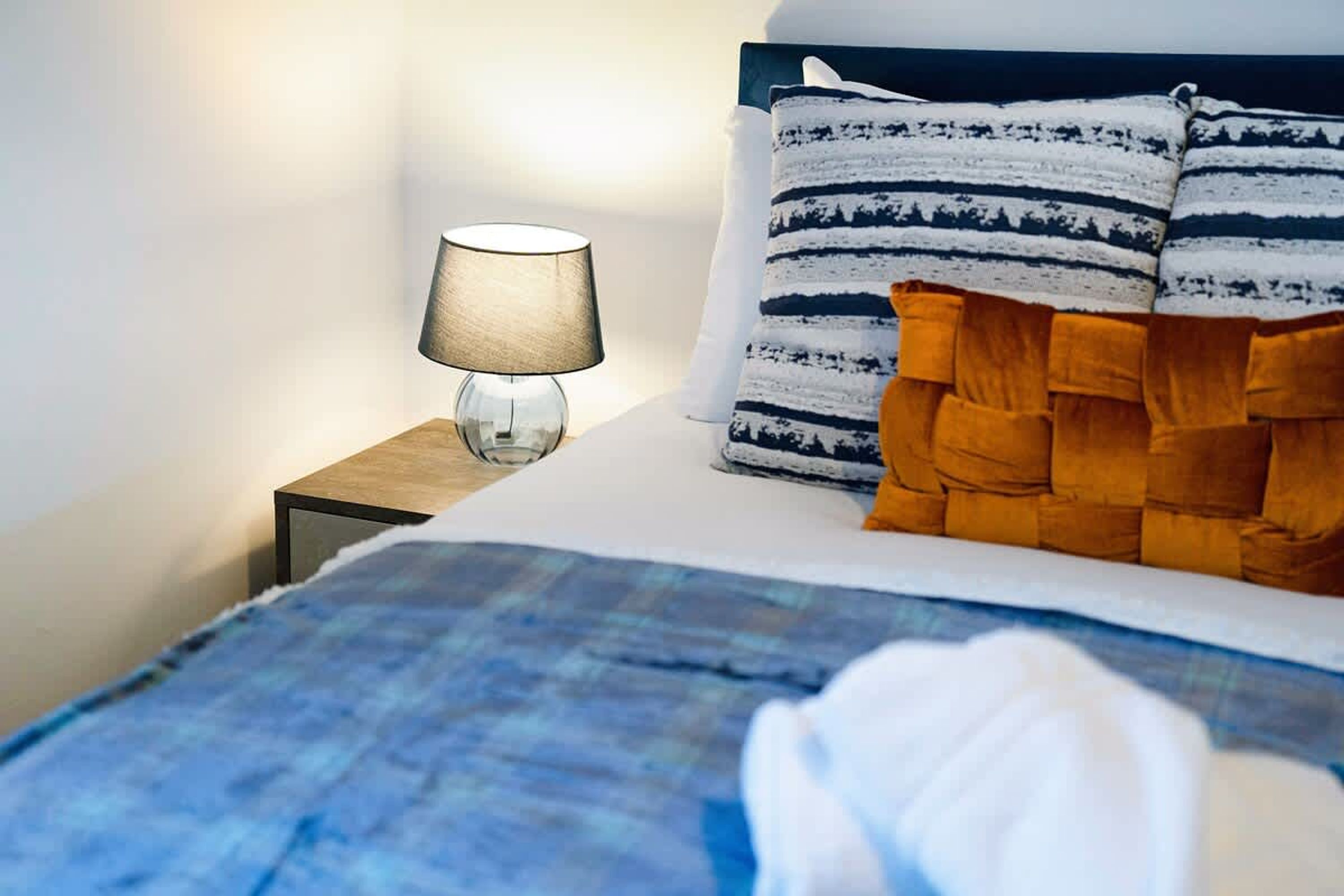 Un lit bien fait, avec une couette à carreaux bleus et blancs, une table de chevet en bois avec une lampe, et une tête de lit agrémentée d’oreillers à rayures.