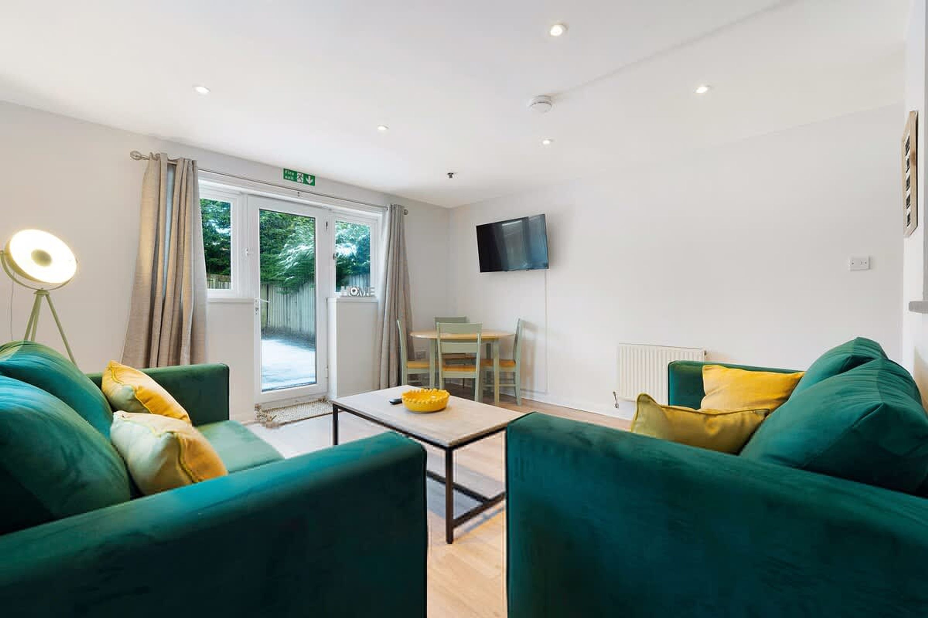 A hotel room with green sofas, a small table, a flat-screen TV, and a window with curtains.
