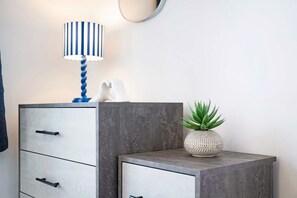 Une chambre moderne avec un meuble gris, une commode blanche, une lampe à rayures bleues et blanches, et une petite plante en pot.
