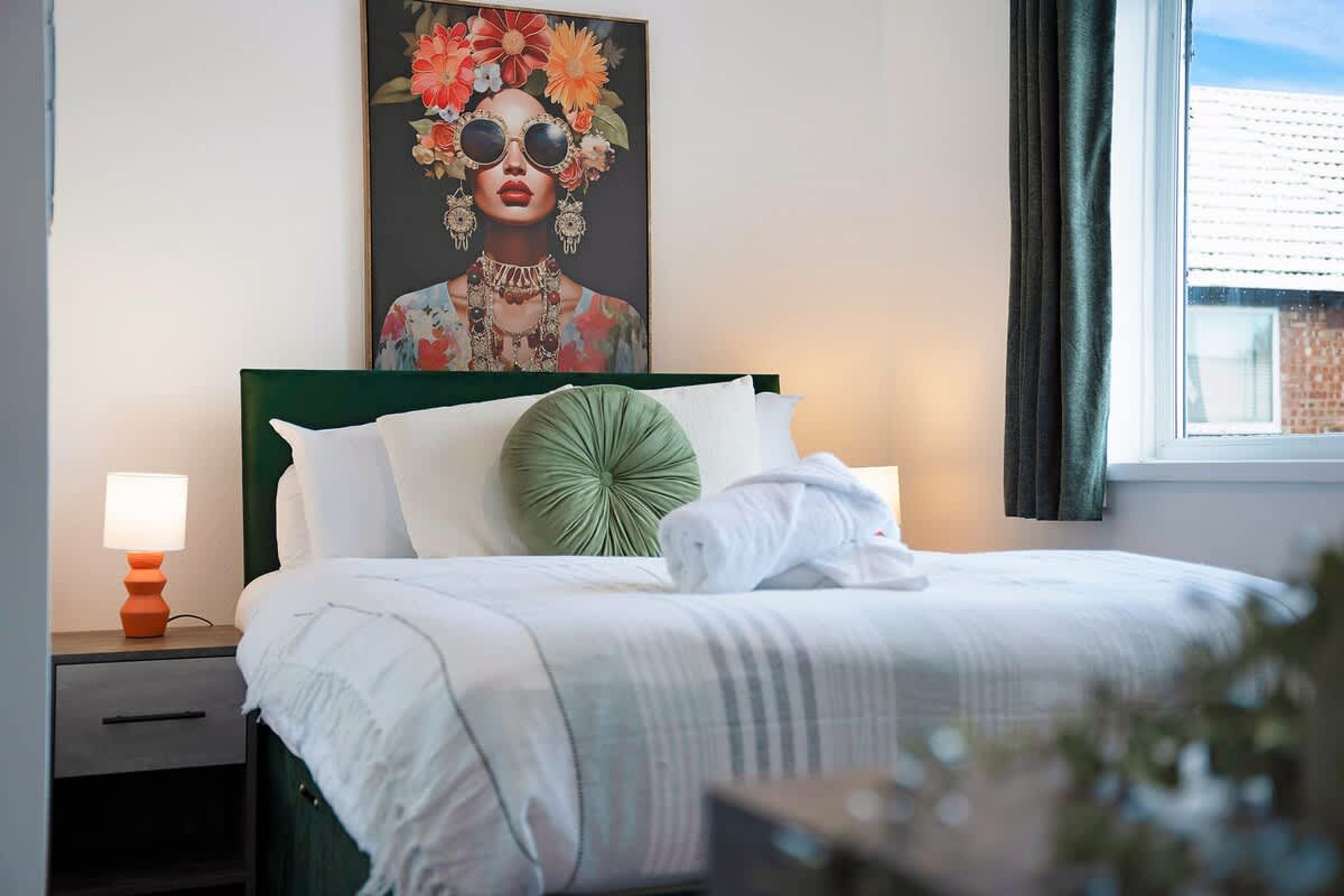 A neatly made bed with white bedding, a green accent pillow, and a framed artwork above it.