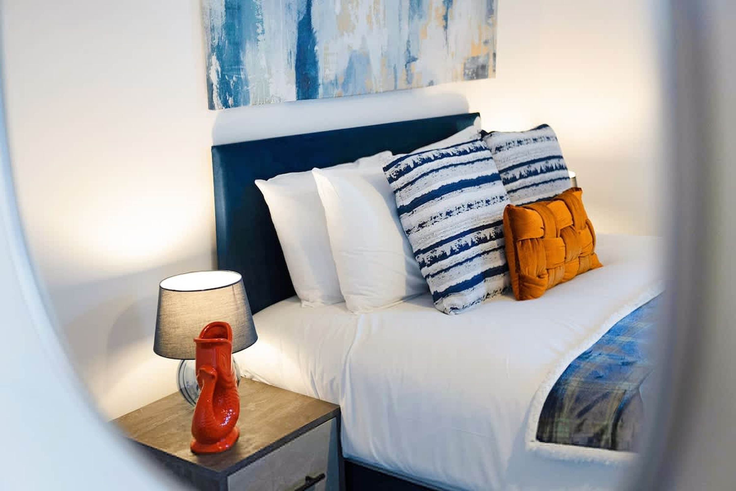 Una cama bien hecha con ropa blanca y almohadas a rayas, una mesita de noche de madera con un objeto rojo encima y una pared con arte abstracto.