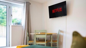 Una habitació amb televisió penjada a la paret que mostra 'NETFLIX', una finestra amb cortines, una taula amb un objecte groc i un sofà verd.