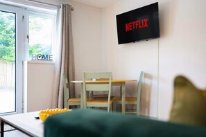 Ein Zimmer mit an der Wand montiertem Fernseher, der 'NETFLIX' anzeigt, einem Fenster mit Vorhängen, einem Tisch mit einem gelben Gegenstand und einem grünen Sofa.