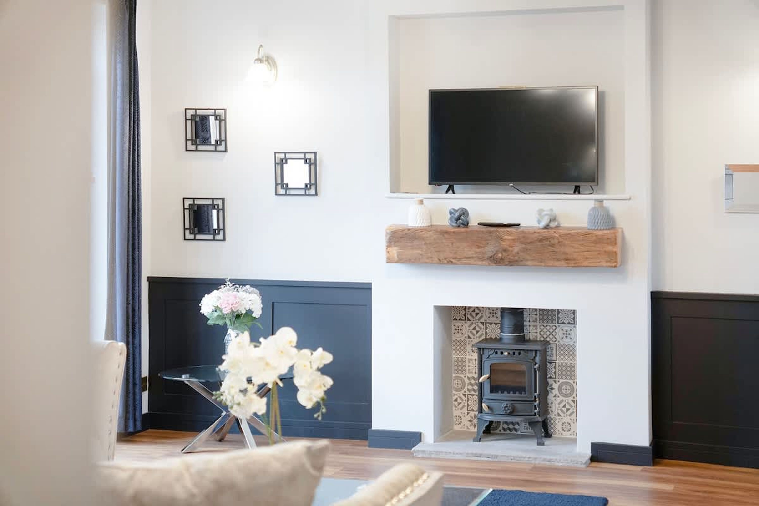 A living room with a flat-screen TV mounted above a fireplace, a wooden shelf with decorative items, and a floral arrangement on a side table.