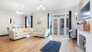 Amplia sala de estar con un sofá blanco, una mesa de centro de vidrio y un tapete azul. Se observan dos lámparas colgantes, una chimenea y un gran ventanal corredero que da a un área exterior.