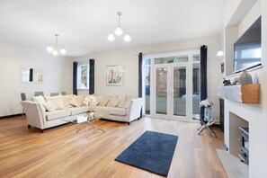 Ein großzügiges Wohnzimmer mit einem weißen Sofa, einem Glastisch und einem blauen Teppich. Zwei Pendelleuchten, ein Kamin und eine große Glasschiebetür, die zu einem Außenbereich führen, sind ebenfalls vorhanden.