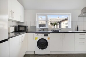 Et moderne køkken med hvide skabe, en mikrobølgeovn, et brødskrin, en teproducent og en vaskemaskine.