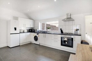A modern kitchen with white cabinetry, a built-in washing machine, a gas stove, and a view of the city through a window. - Stylish 2BR House in Ashington by Stay With Us (Ashington)