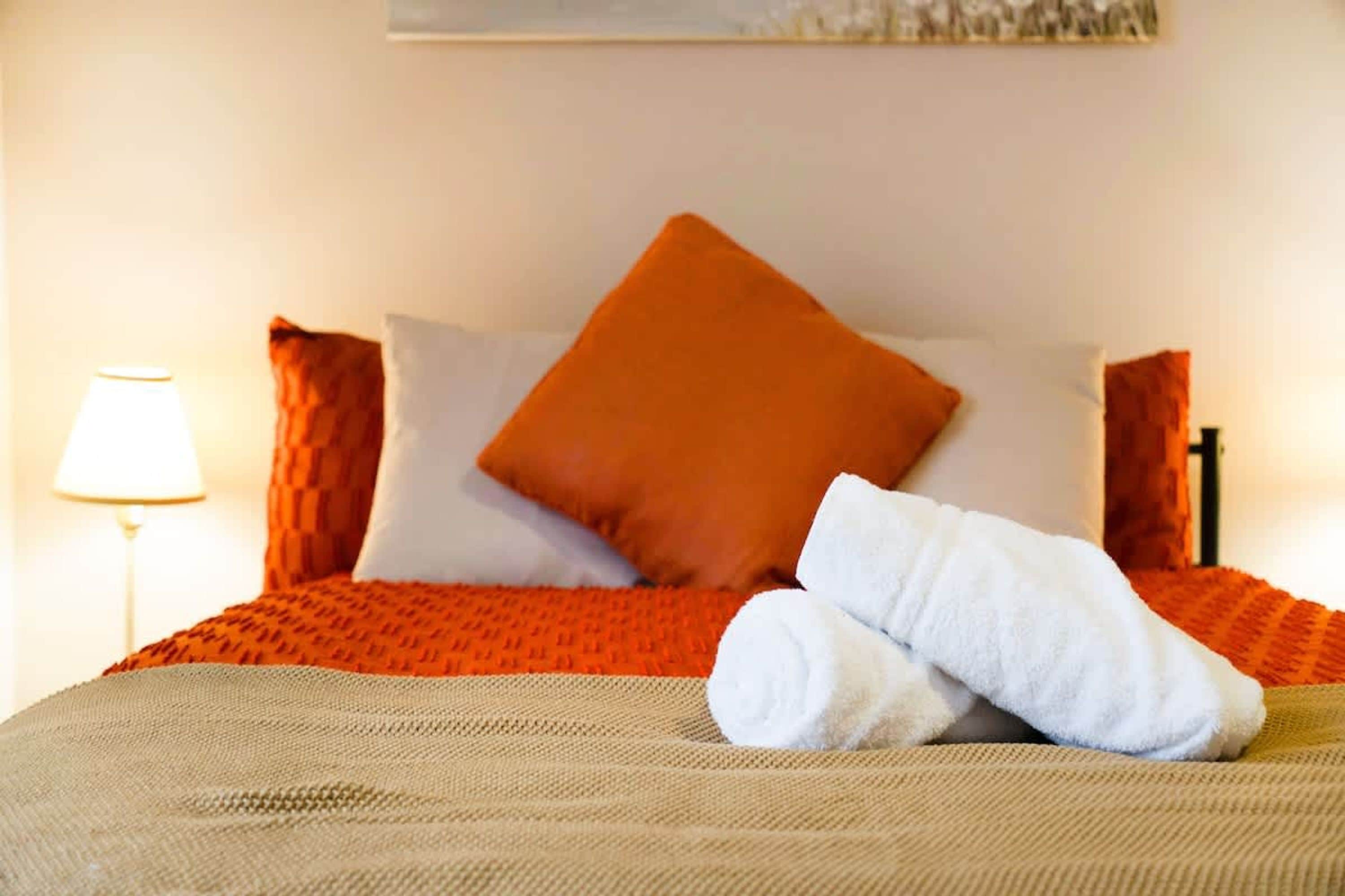 En pænt redt seng med et brunt sengetæppe, to hvide puder og en orange detaljepude.