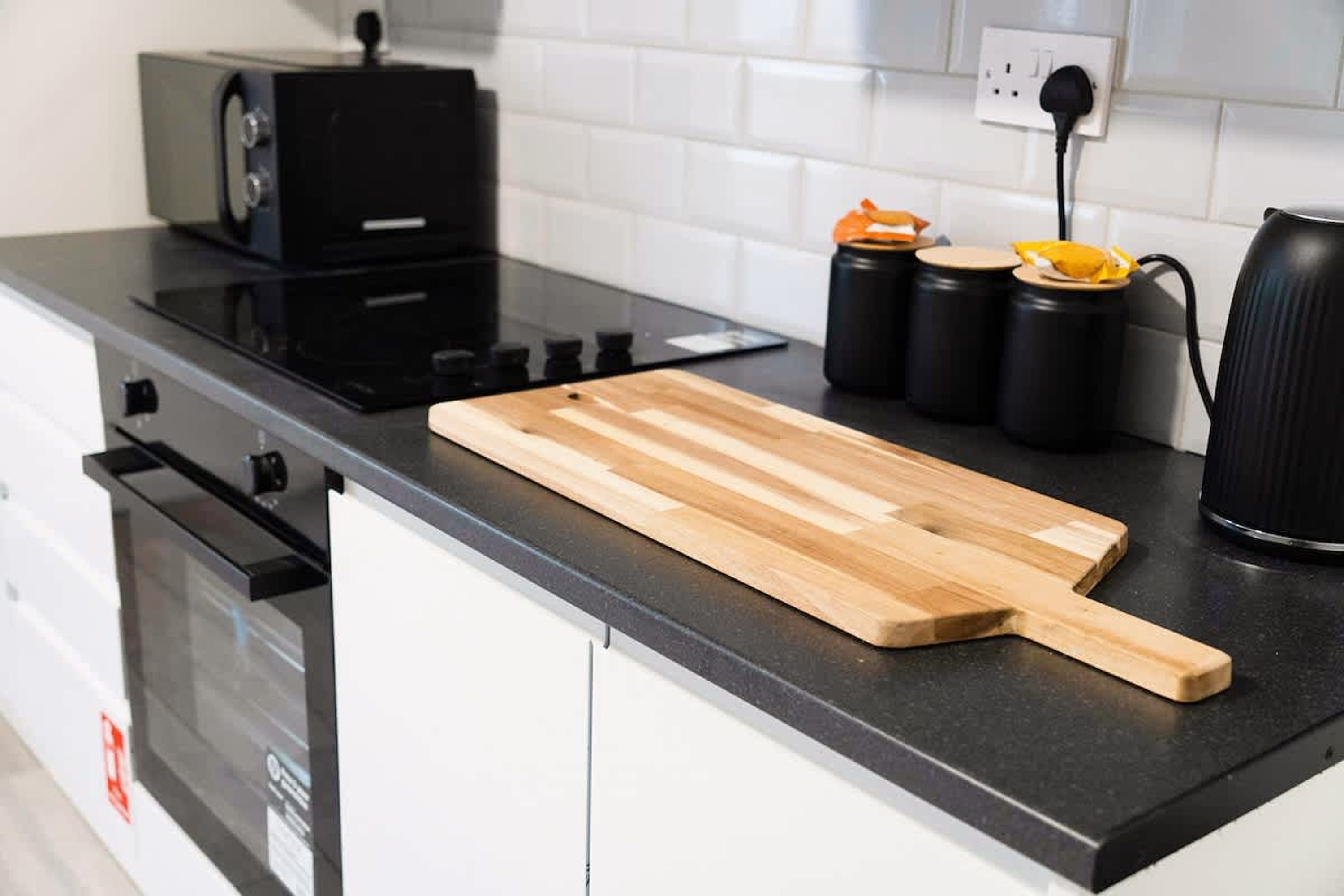 A kitchen with a black countertop, a wooden cutting board, a black kettle, and two black containers with orange lids.