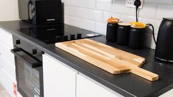 A kitchen with a black countertop, a wooden cutting board, a black kettle, and two black containers with orange lids.