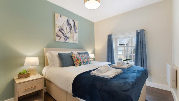 A hotel room with a bed, bedside table, lamp, artwork, and a window with curtains. - Chic Flat Steps From Barnard Castle High Street (Barnard Castle)