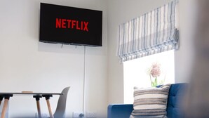 A living room with a TV mounted on the wall displaying 'NETFLIX,' a laptop on a table, and a sofa with a striped cushion. - Chic Flat Steps From Barnard Castle High Street (Barnard Castle)