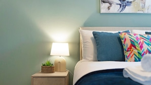 Una cama bien hecha con un almohadón azul y de varios colores, una almohada blanca y una manta azul. Hay una mesita de noche con una lámpara y una planta.