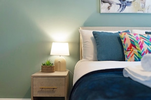 A neatly made bed with a blue and multicolored pillow, a white pillow, and a blue blanket. There is a small bedside table with a lamp and a plant. - Chic Flat Steps From Barnard Castle High Street (Barnard Castle)