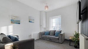 A modern living room with a grey sofa, a smaller grey armchair, and a coffee table. There are two wall-mounted paintings, a window with blinds, and a radiator. - Stockton 5BR for Contractors With Free Parking (Stockton-on-Tees)