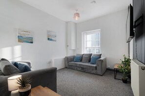A modern living room with a grey sofa, a smaller grey armchair, and a coffee table. There are two wall-mounted paintings, a window with blinds, and a radiator. - Stockton 5BR for Contractors With Free Parking (Stockton-on-Tees)