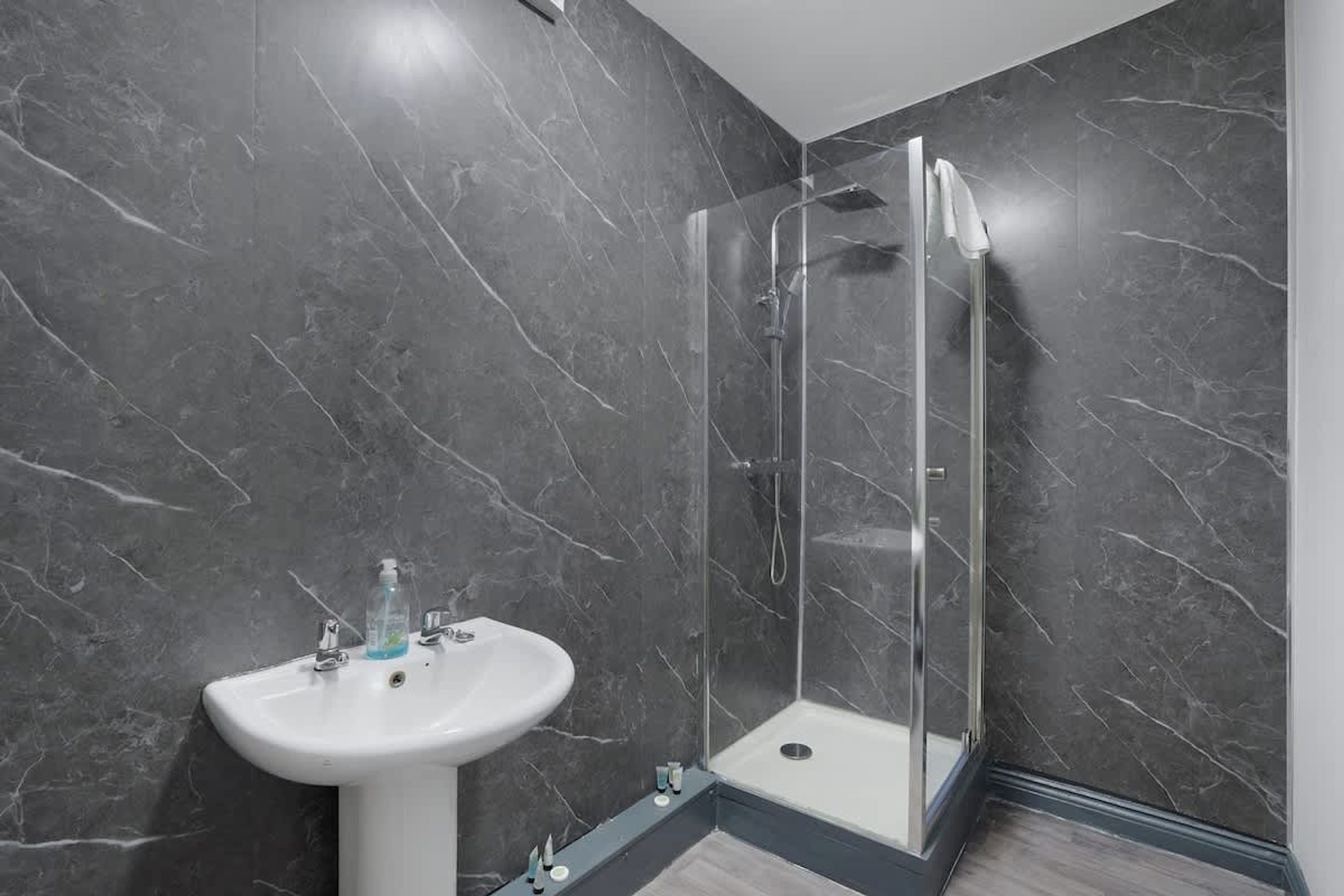 Ein Badezimmer mit einem weißen Standwaschbecken, einer Duschkabine aus Glas und dunkel marmorierten Wänden.