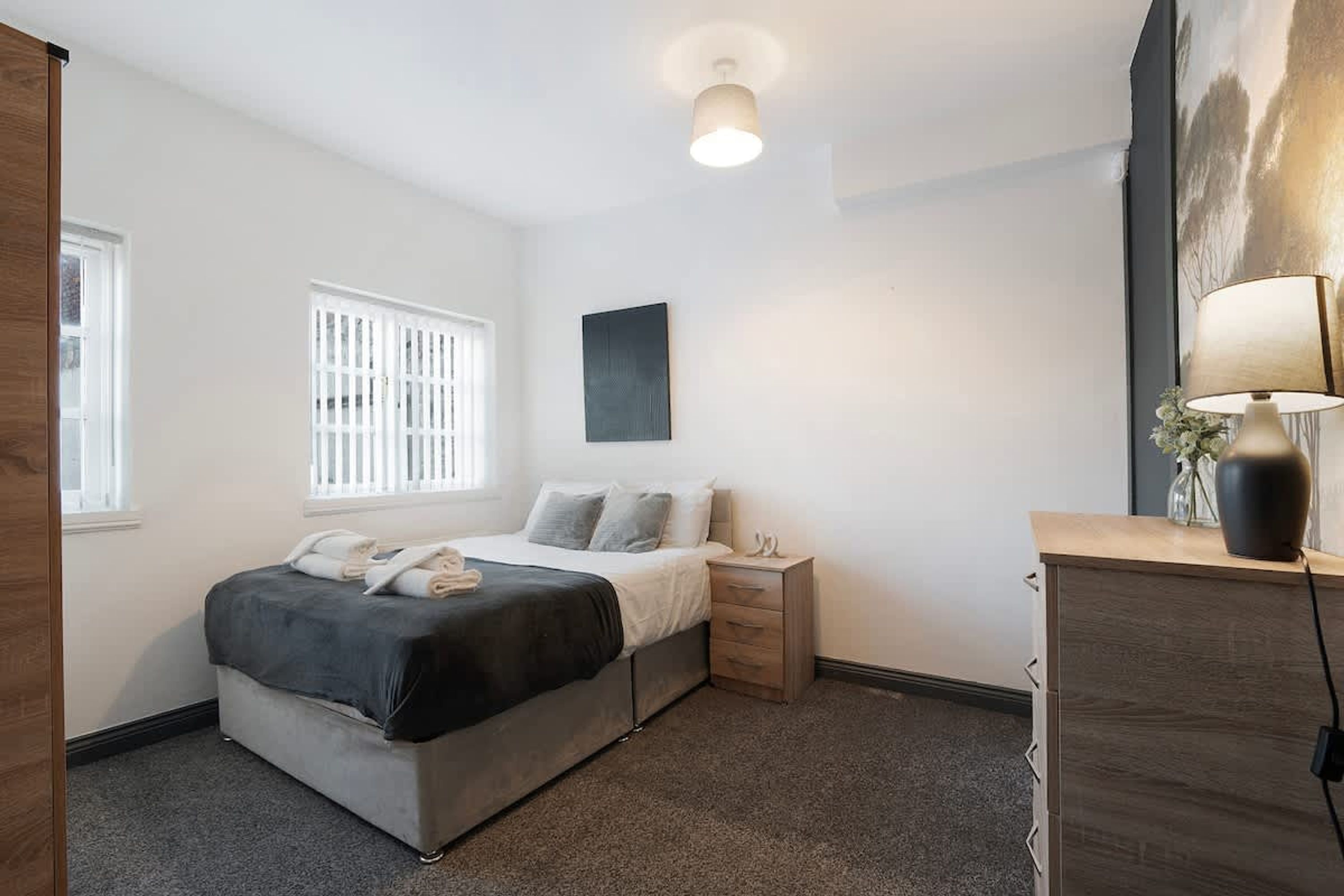 A bedroom with a bed, bedside table, lamp, and a window with blinds.