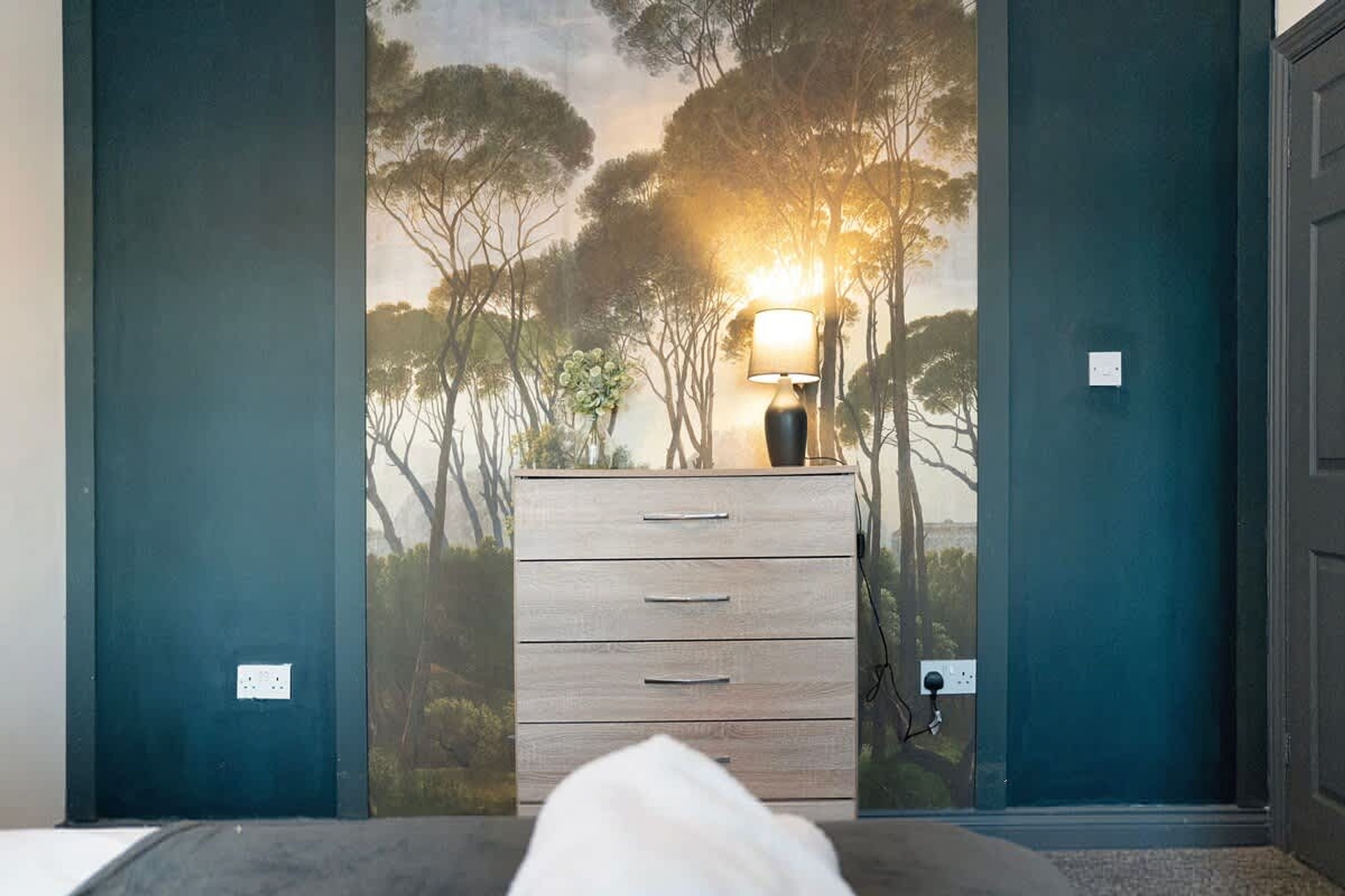 Et værelse med et tapet der viser et skovmotiv, et natbord med en lampe, og en dør med en stikkontakt.