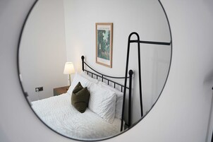 Una cama con ropa de cama blanca y una almohada verde, una lámpara de noche y una ilustración botánica enmarcada en la pared.