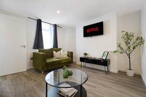 Una sala de estar moderna con un sofá verde, una mesa de centro de vidrio, una consola negra con objetos decorativos, una planta en maceta y un televisor montado en la pared mostrando 'NETFLIX'.