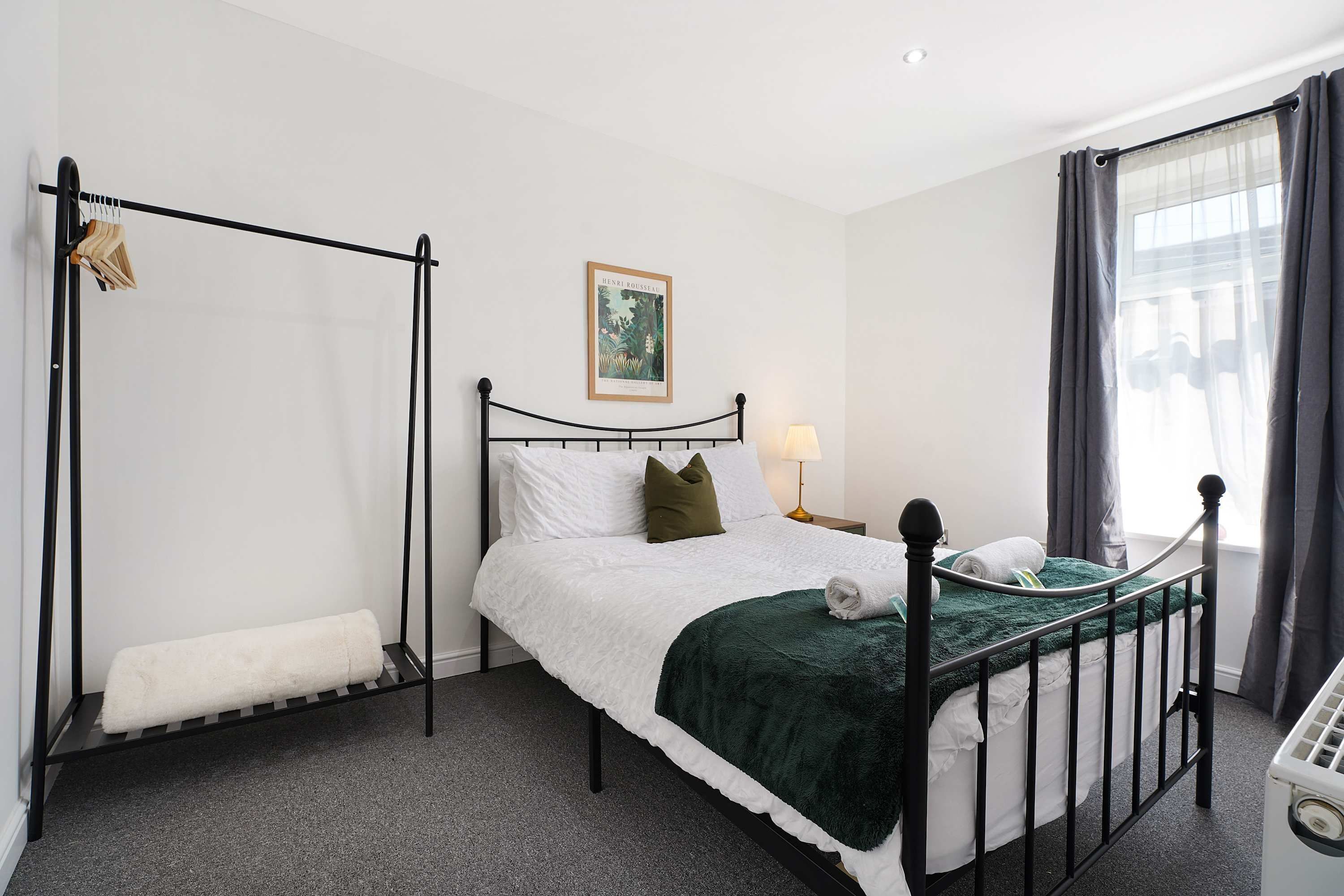 A single bed with a headboard, a bedside table with a lamp, a window with curtains, and a wall-mounted radiator.