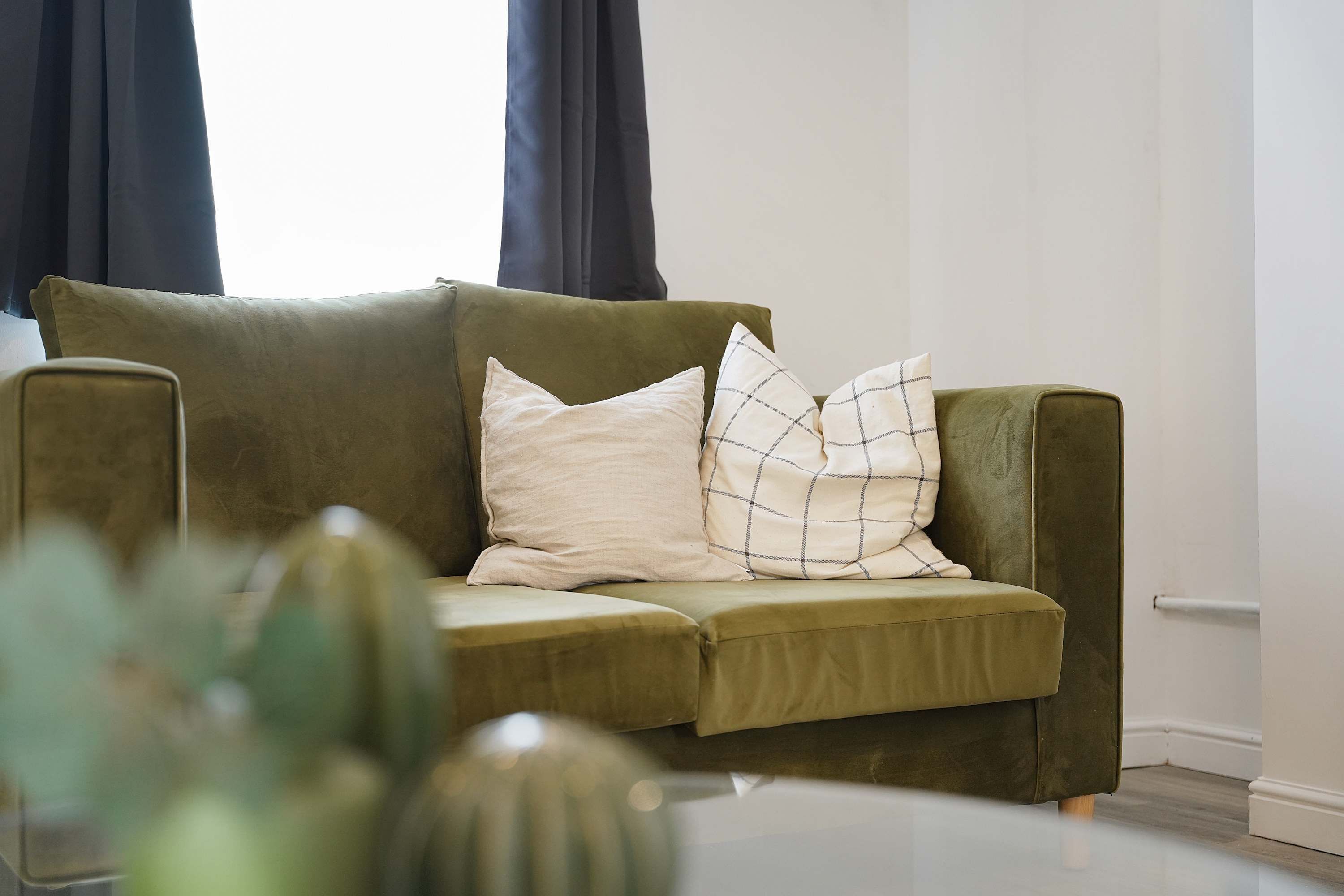 A green sofa with two pillows, a white table with a vase, and a window with curtains.