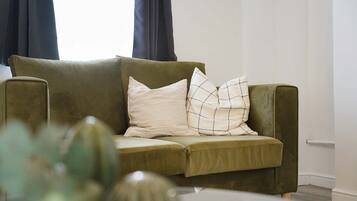 A green sofa with two pillows, a white table with a vase, and a window with curtains.